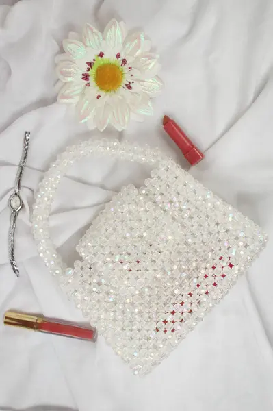 Handmade white purse made with bright beads on gray fabric with makeup tool and fake daisy flower. Top view.