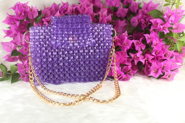 Handmade purple purse made with bright beads on white furry blanket with pink bougainvillea flower. Purple purse with gold handle.