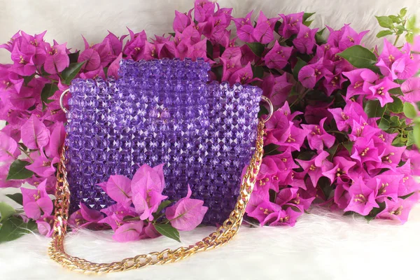 Handmade purple purse made with bright beads on white furry blanket with pink bougainvillea flower. Purple purse with gold handle.