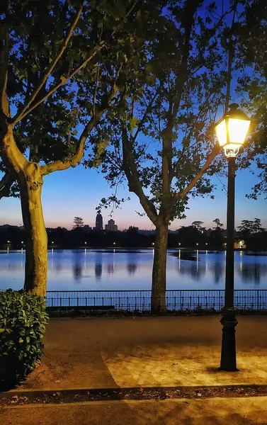 Madrid ufuk çizgisi şafakta, Casa de Campo gölünden görünüyor, ön planda yanan bir sokak lambasıyla..