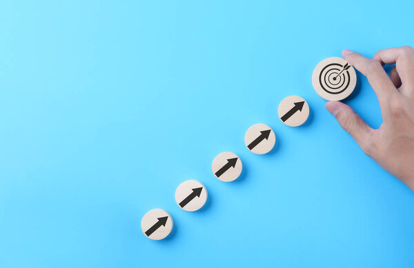 Business achievement goal and objective target concept. Businessman hand putting virtual business target and arrow which screen on round wooden block. Growth investing, Blue background, Copy space,
