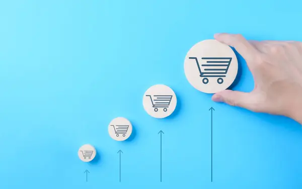 Online sale business growth concept. Hand holding wooden block with bigger shopping trolley cart icon on blue background. Increasing retail trend, Commerce shop, Marketing strategies to Increase sale,