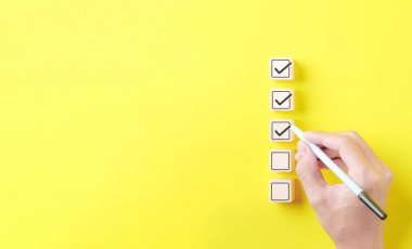 Checklist concept. Person using a black ink pen to correct sign and tick check mark on wooden block with copy space for the checklist on yellow background. Business survey questionnaire, exam process, clipart