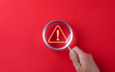 Uyarı sistemi konsepti. Bir el, parlak bir ünlem işaretinin üzerinde kırmızı bir zeminde bir büyüteç tutar. Bilgisayar ağına, suç ve virüse, kötü niyetli yazılıma sızıldı,