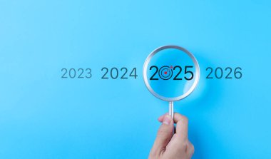 2025 Yılbaşı İş Hedefleri Konsepti. 2025 yılına ait bir büyüteçle el ele tutuşmak, iş hedefleri ve gelecekteki hedeflere odaklanmayı temsil eden bir hedef simgesiyle birlikte bir büyüteçtir. Pazarlama eğilimi