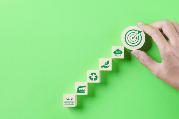 Green Energy and Sustainable Concept. Hand holding a wooden block a target with a leaf, with green energy and sustainability icons on a green background. Carbon footprint, global warming, net zero,
