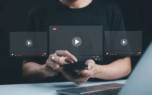Canlı yayın ve video içeriği oluşturma kavramı. Bir adam, canlı yayın, dijital medya ve video içeriği oluşturmayı sembolize eden, şeffaf video oynatıcı arayüzleri olan bir akıllı telefon aygıtı kullanıyor.