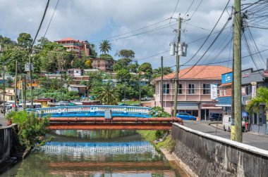 Castries 'deki sokak sahnesi, Saint Lucia' nın başkenti, kanal, mavi köprü, kırmızı borular, yamaçlardaki evler, tropikal yeşillikler, palmiye ağaçları, elektrik direkleri, canlı Karayip mimarisi Şubat 2025