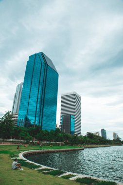 Bangkok, Tayland. 4 Temmuz 2022. Benchakitti Parkı 'nın Cityscape' inde gündüz vakti göl kenarında dinlenen insanlar. Arka planında gökdelenler olan bir iş merkezi. dikey çekim