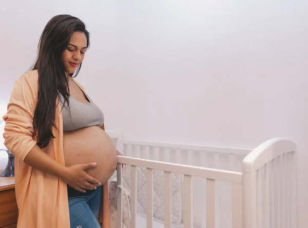 妊娠中のラテン系の女性彼女の腹に触れる彼女の赤ちゃんの白いベビーベッドの横に — ストック写真