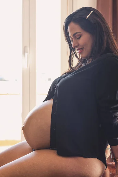 若いです妊娠中の女性 座っています彼女の腹を見て後ろに窓と黒のブラウス — ストック写真