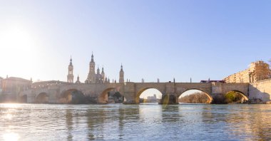 Arka planda taş köprü ve Pilar Katedrali, sol tarafında yakıcı bir güneş ve Ebro nehrinin suyunda yansıma var.