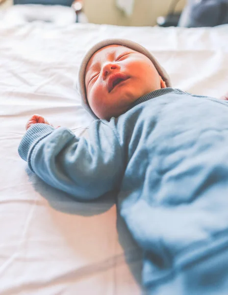ベッドの中で新生児の睡眠は 彼の左腕を持ち上げた 青い服とキャップを身に着けて — ストック写真
