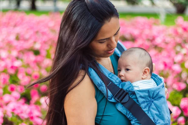 Latina mom and her baby in a blue baby carrier, walking through a park with pink flowers.