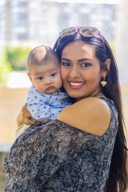 Mutlu anne parkta bebeğini kucaklıyor, gülümsüyor ve kameraya bakıyor..