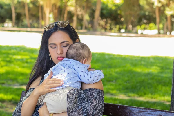 Young Latina mom with her eyes closed, holding her baby in a park