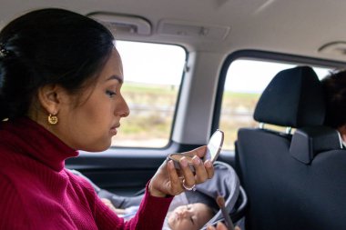 Latin woman putting on makeup in a car with her baby next to her clipart