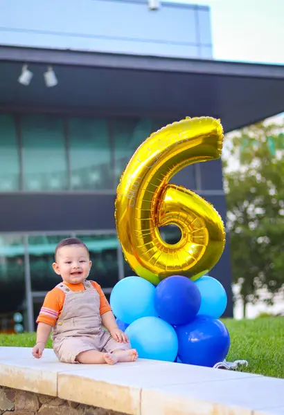 Bebek gülümsüyor, mutlu ve arka planda 6 numaralı balonla parkta oturuyor.