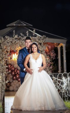 Neşeli bir çift düğünlerinde kucaklaşıp, yumuşak ışıklandırma ve romantik dekor arasında aşklarını sergiliyor. Arkaplanda çiçek aranjmanları ve balonlar yer alıyor, büyülü bir akşam atmosferini yakalıyorlar..