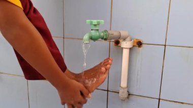 Mavi seramik duvarlı yeşil bir musluktan ayaklarını suda yıkayan bir çocuk.