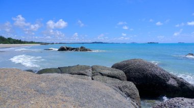 Ön planda büyük, pürüzsüz kayalar olan tropikal bir plajın çarpıcı panoramik görüntüsü. Turkuaz su yavaşça kıyıya vurur ve beyaz kumlu sahil mesafeye kadar uzanır.