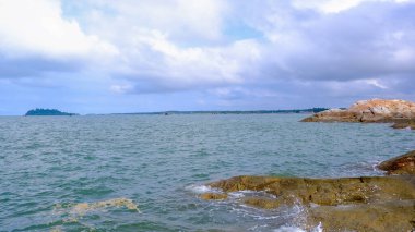 Kayalık kıyı şeridi ve küçük adaları olan güzel bir deniz manzarası. Gökyüzü parçalı bulutludur, ve su dalgaların açık mavi ve beyaz karışımıdır..