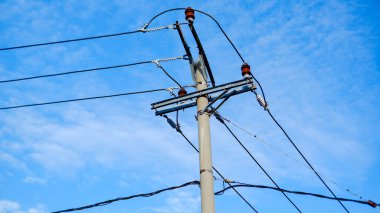 Parlak mavi gökyüzü boyunca uzanan kablolu beton bir elektrik direğinin fotoğrafı. Turuncu yalıtkanları dışarı fırlayan güçlü direk, zarafetle karşı karşıya gelen bir dizi elektrik hattını destekler. 