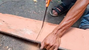 Mini testereyle turuncu bir tahtayı kesmeye çalışan bir adamın eli. Sağ el testereyi tutuyor ve sol el tahtayı tutuyor böylece kesilirken sallanmıyor.