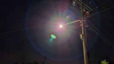 Bir elektrik direğine asılmış bir sokak lambasının açık bir gece fotoğrafı. Yoğun parlaklıkla karanlık gökyüzünü aydınlatıyor..