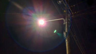 Bir elektrik direğine asılmış bir sokak lambasının açık bir gece fotoğrafı. Yoğun parlaklıkla karanlık gökyüzünü aydınlatıyor..