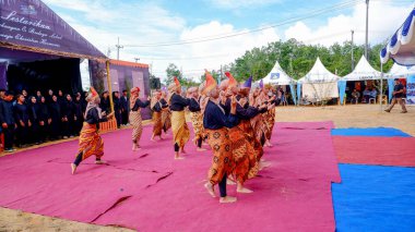 MUNTOK, INDONEZYA - 27 YIL 2024: Endonezyalı geniş bir grup geleneksel dansçıyı canlı kırmızı ve mavi bir halının üzerinde dans ederken gösteren geniş, dinamik bir çekim