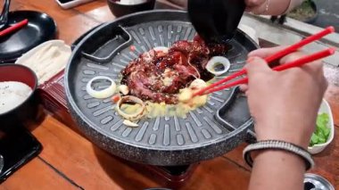 Sıcak Kore barbekü ızgarasında marine edilmiş et ve sebze pişiren insanların görüntüleri. Yemek çubuğu ve maşa kullanan eller, yan yemekler, pirinç ve taze sebzelerle çevrili yemekleri hazırlar.