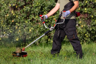 Worker in protective clothing with electric mower in his hands mows the grass on the lawn.