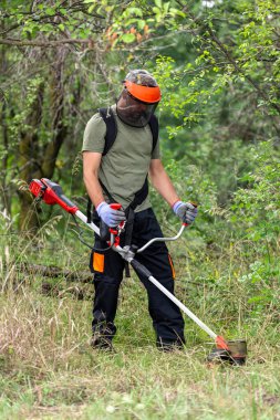 Young adult man in protective clothes mowing green grass with electrical lawn trimmer at garden. Side view.