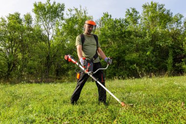 Man mowing the grass at his garden by using string trimmer with protective helmet and gloves.