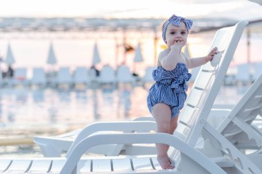 Baby girl on summer vacation, standing on lounger next to swimming pool. Sunset in the background.