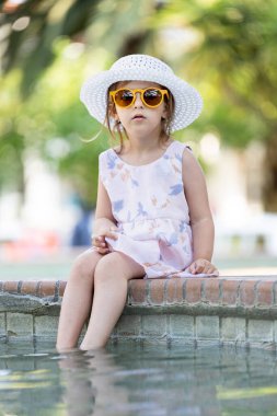 Güneş gözlüklü, güneş şapkalı, havuzun kenarında oturan şık bir kız..