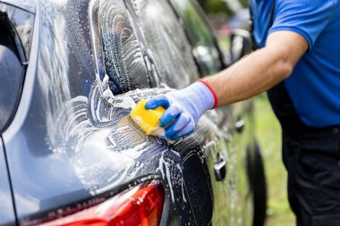 Adam dışarıda sarı süngerle araba yıkıyor.