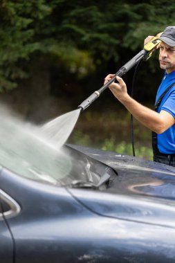 Adam arka bahçede su tazyikli çamaşır makinesiyle arabasını yıkıyor.