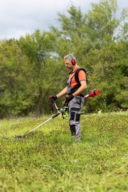 Bulutlu bir yaz gününde, örtülü erkek çiftçi bahçede ot kesiciyle çim biçiyor. Bahçe konsepti.