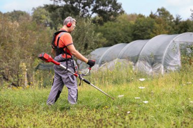 Koruyucu giysi ve eldiven giyen işçilerin doğa arka planında çim biçen bir çim biçme makinesi kullanırken açık hava görüntüsü