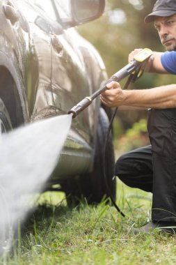 Koruyucu giyimli profesyonel bir adam arka bahçede arabasını yıkamak için su tazyikli çamaşır makinesi kullanıyor.