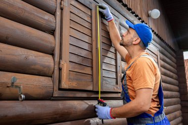 Professional using measure tape for window shutter replacement on wooden house. Home reparation concept.