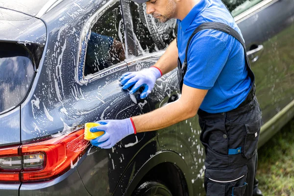 Adam dışarıda sarı süngerle araba yıkıyor.