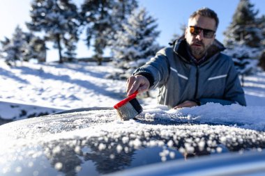 Modern adam fırçayı tutarken güneşli kış gününde arabanın çatısından karı temizlerken.