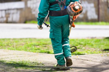 Yeşil üniformalı bir işçi, çim biçme makinesi taşır. Açık hava bakım işlerinde ekipmanlara odaklanır.