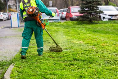Yüksek görüş mesafesi olan bir işçi, kamusal alanların bakımını üstleniyor, sokak boyunca uzanan çimenleri biçmek için bir tel budayıcı ustalıkla manevra yapıyor.