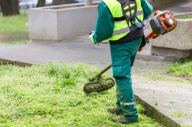 Yeşil üniformalı ve koruyucu kıyafetli bir işçi kaldırımın yanındaki çimenleri biçmek için çim biçme makinesi kullanıyor.