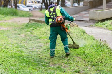 Yeşil üniformalı ve koruyucu kıyafetli bir işçi kaldırımın yanındaki çimenleri biçmek için çim biçme makinesi kullanıyor.