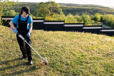 İş üniformalı bir kadın, güneşli bir günde yeşil bir parkta ya da arka bahçede tel budayıcı kullanıyor.